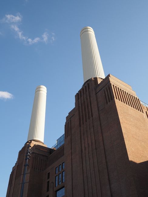 A low-angle view of Battersea Power Station with its distinctive red-brick facade and four tall white chimneys extending into a clear blue sky, featuring minimal clouds. The brickwork shows vertical d