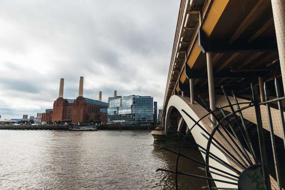 A low-angle view of Battersea Power Station with its distinctive red-brick facade and four tall white chimneys extending into a clear blue sky, featuring minimal clouds. The brickwork shows vertical d