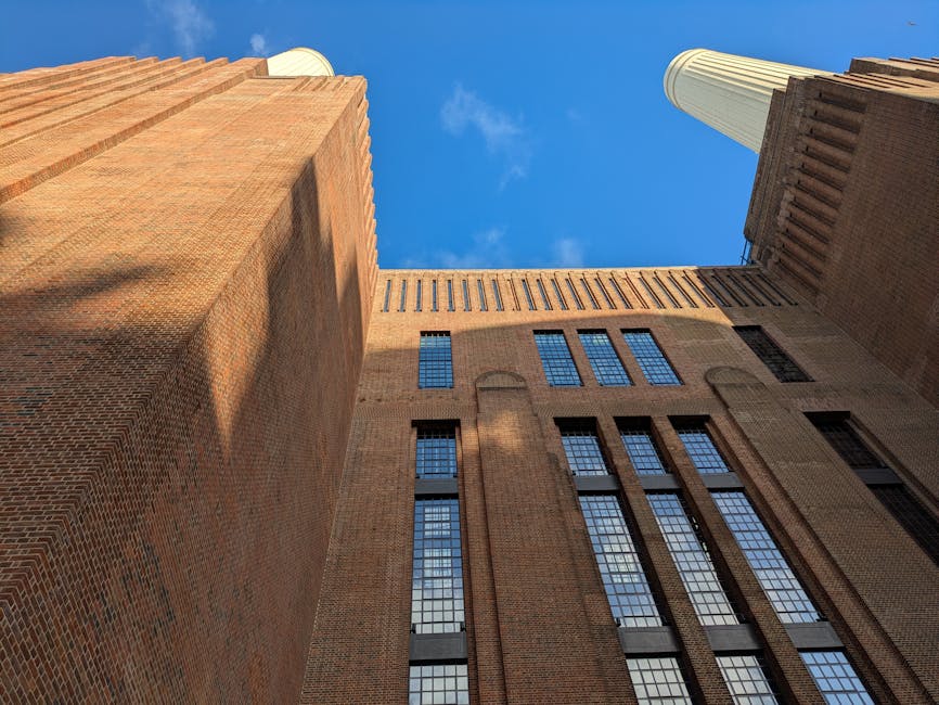 Looking up at towering brick buildings against a bright blue sky with a few wispy clouds, showcasing the architectural details and grid-like pattern of windows on the facades. The scene highlights the